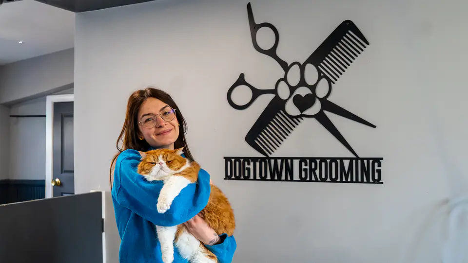 Groomer holding a ginger-and-white cat in front of the Dogtown Grooming sign