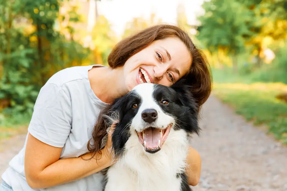 A smiling woman hugs a black and white Border Collie on a sunlit tree-lined path.