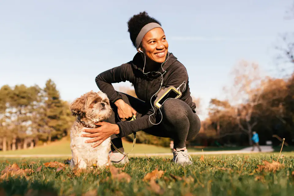 A woman in athletic wear crouches on a park lawn and pets a small shaggy dog.